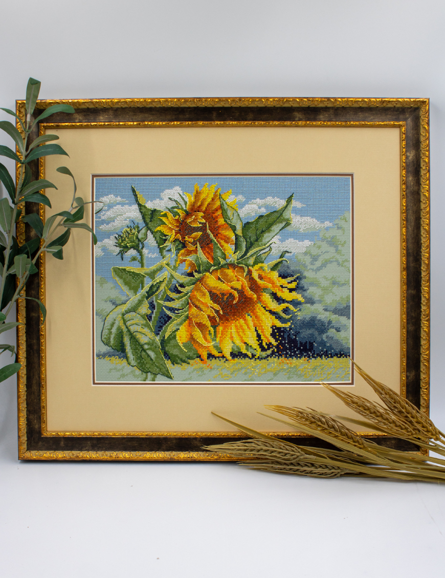 A completed cross-stitch of two large, vibrant yellow and orange sunflowers with textured green leaves, depicted as swaying in the wind against a light blue sky with fluffy white clouds.