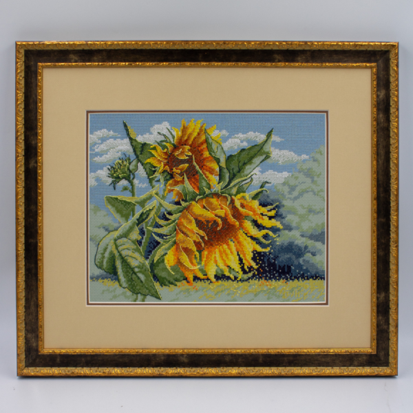 A completed cross-stitch of two large, vibrant yellow and orange sunflowers with textured green leaves, depicted as swaying in the wind against a light blue sky with fluffy white clouds.