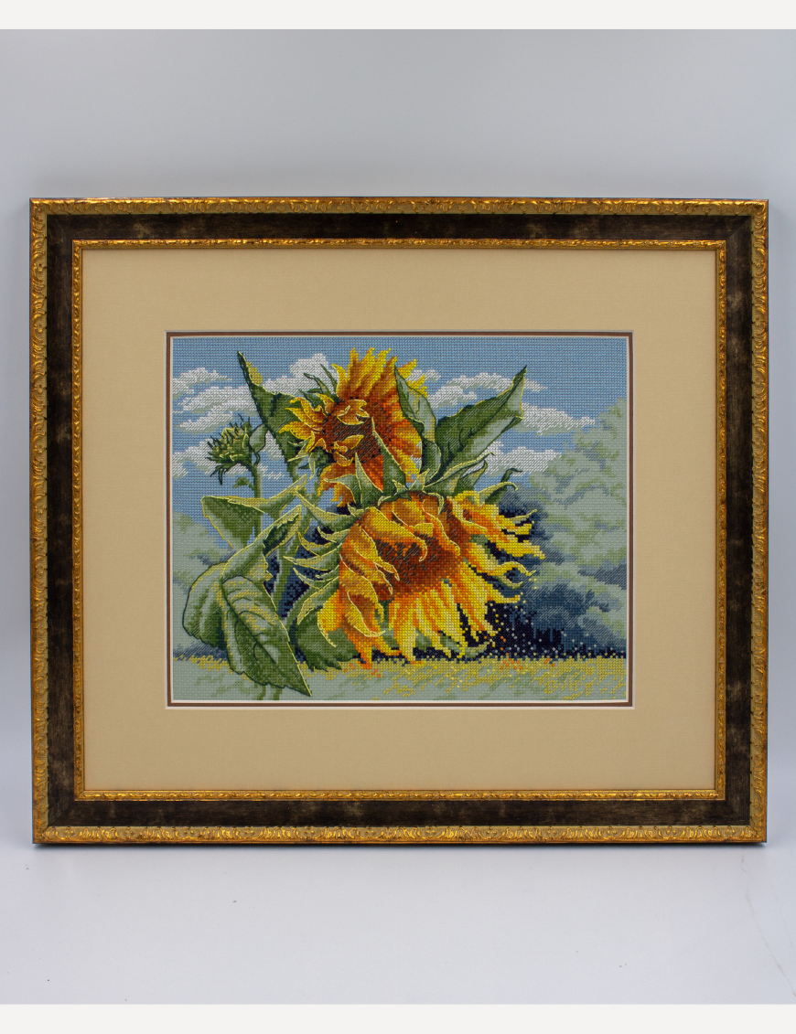 A completed cross-stitch of two large, vibrant yellow and orange sunflowers with textured green leaves, depicted as swaying in the wind against a light blue sky with fluffy white clouds.