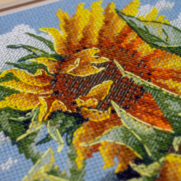 A completed cross-stitch of two large, vibrant yellow and orange sunflowers with textured green leaves, depicted as swaying in the wind against a light blue sky with fluffy white clouds.