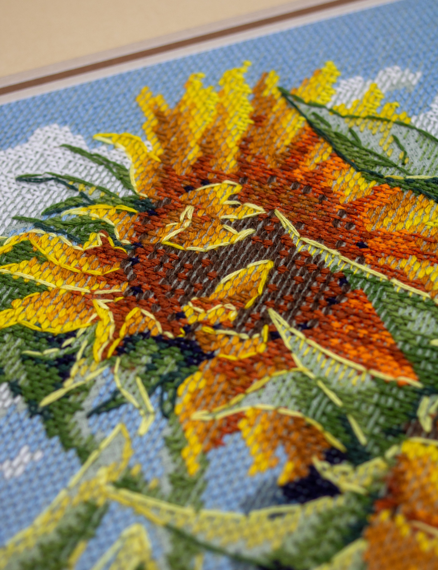 A completed cross-stitch of two large, vibrant yellow and orange sunflowers with textured green leaves, depicted as swaying in the wind against a light blue sky with fluffy white clouds.