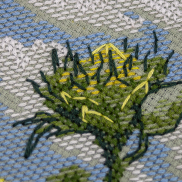 A completed cross-stitch of two large, vibrant yellow and orange sunflowers with textured green leaves, depicted as swaying in the wind against a light blue sky with fluffy white clouds.
