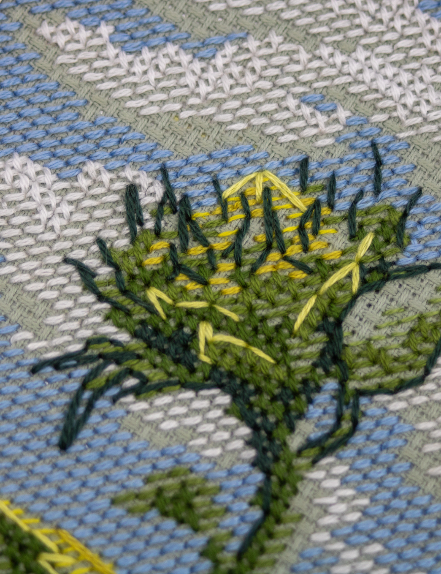 A completed cross-stitch of two large, vibrant yellow and orange sunflowers with textured green leaves, depicted as swaying in the wind against a light blue sky with fluffy white clouds.