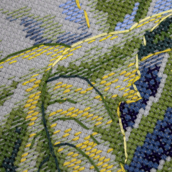 A completed cross-stitch of two large, vibrant yellow and orange sunflowers with textured green leaves, depicted as swaying in the wind against a light blue sky with fluffy white clouds.