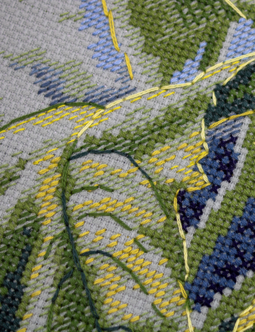 A completed cross-stitch of two large, vibrant yellow and orange sunflowers with textured green leaves, depicted as swaying in the wind against a light blue sky with fluffy white clouds.