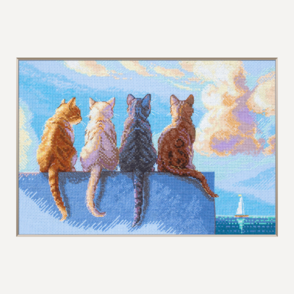 A completed cross-stitch landscape showing four cats (orange, cream, dark grey, and brown) sitting on a blue wall with their backs to the viewer. They are looking out over a blue ocean with a small white sailboat, under a sky filled with fluffy yellow and pink clouds.
