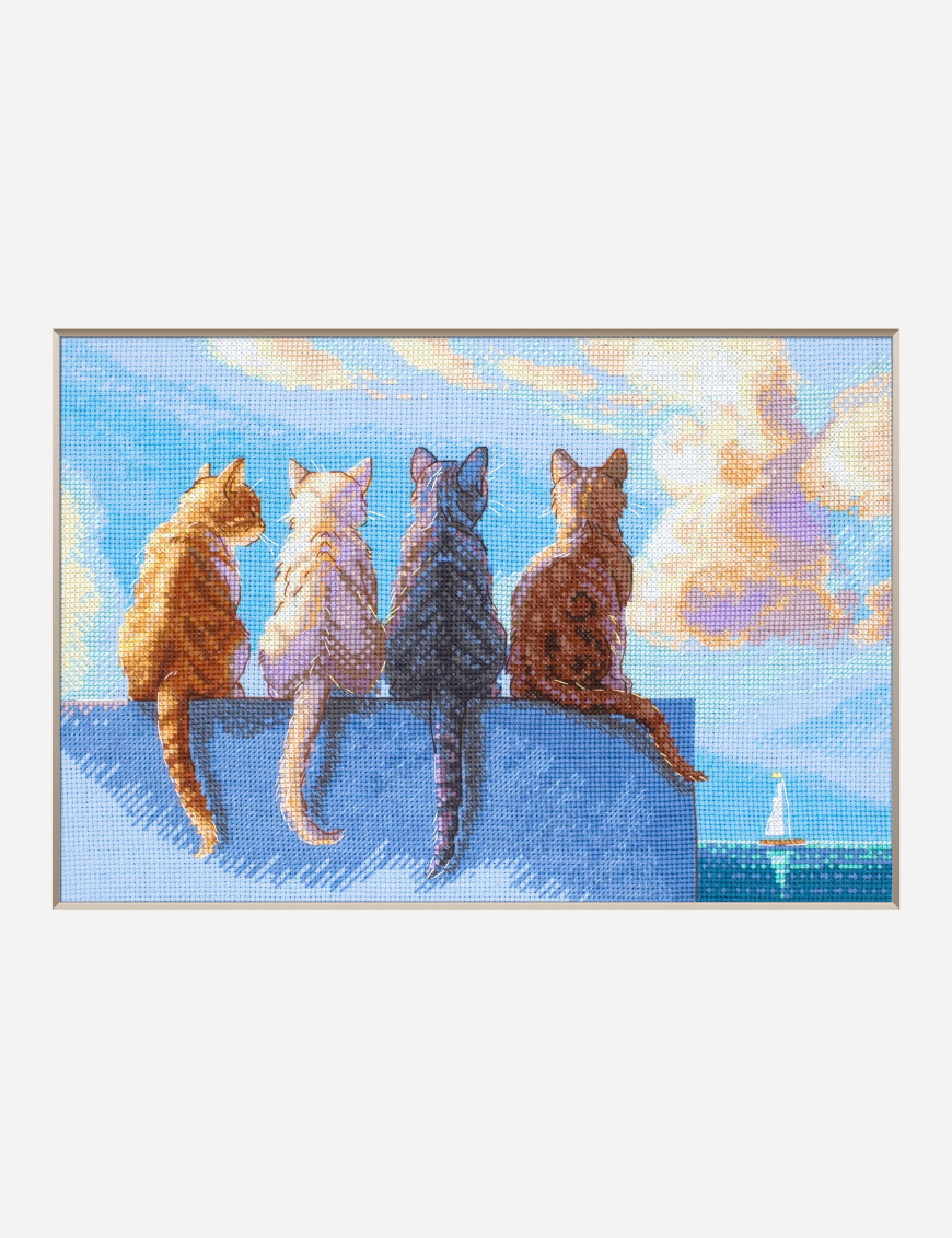 A completed cross-stitch landscape showing four cats (orange, cream, dark grey, and brown) sitting on a blue wall with their backs to the viewer. They are looking out over a blue ocean with a small white sailboat, under a sky filled with fluffy yellow and pink clouds.
