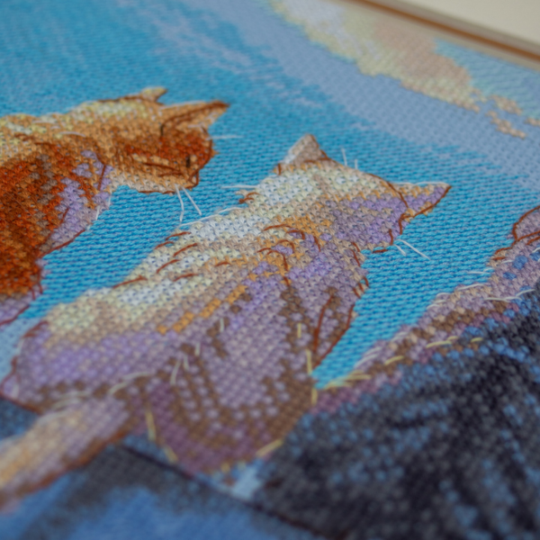 A completed cross-stitch landscape showing four cats (orange, cream, dark grey, and brown) sitting on a blue wall with their backs to the viewer. They are looking out over a blue ocean with a small white sailboat, under a sky filled with fluffy yellow and pink clouds.