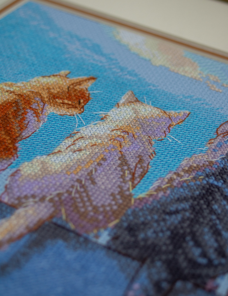 A completed cross-stitch landscape showing four cats (orange, cream, dark grey, and brown) sitting on a blue wall with their backs to the viewer. They are looking out over a blue ocean with a small white sailboat, under a sky filled with fluffy yellow and pink clouds.