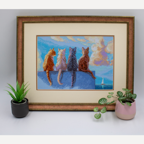 A completed cross-stitch landscape showing four cats (orange, cream, dark grey, and brown) sitting on a blue wall with their backs to the viewer. They are looking out over a blue ocean with a small white sailboat, under a sky filled with fluffy yellow and pink clouds.