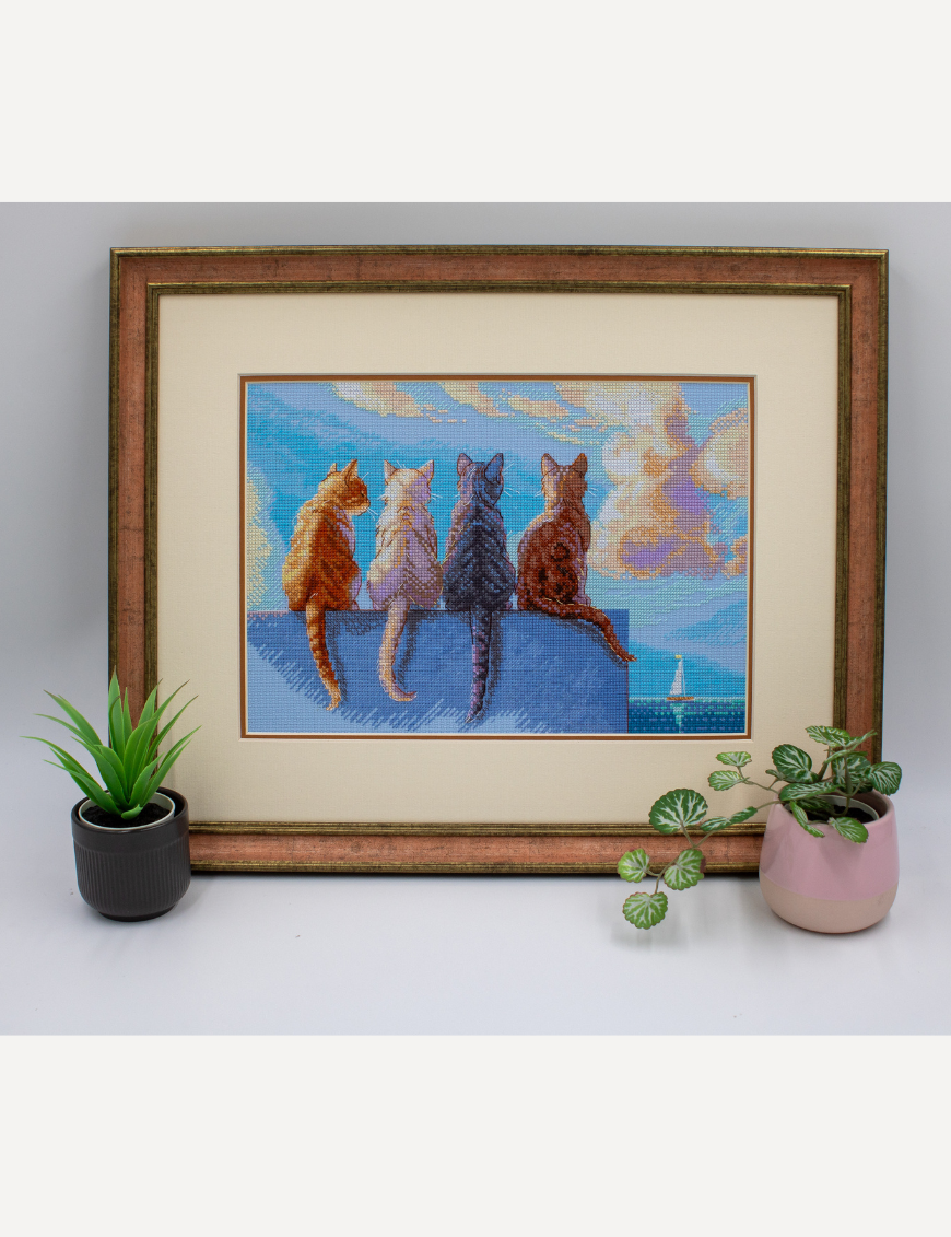A completed cross-stitch landscape showing four cats (orange, cream, dark grey, and brown) sitting on a blue wall with their backs to the viewer. They are looking out over a blue ocean with a small white sailboat, under a sky filled with fluffy yellow and pink clouds.