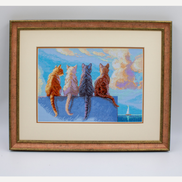 A completed cross-stitch landscape showing four cats (orange, cream, dark grey, and brown) sitting on a blue wall with their backs to the viewer. They are looking out over a blue ocean with a small white sailboat, under a sky filled with fluffy yellow and pink clouds.