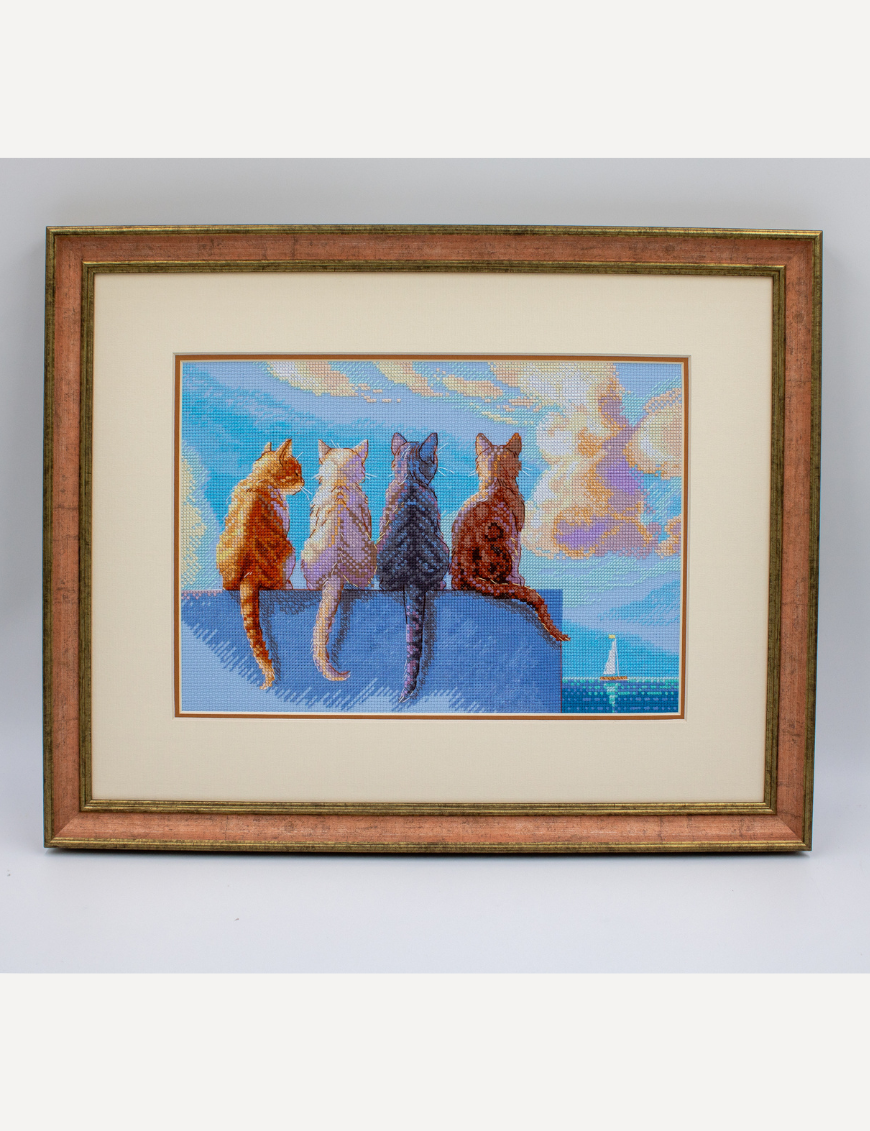 A completed cross-stitch landscape showing four cats (orange, cream, dark grey, and brown) sitting on a blue wall with their backs to the viewer. They are looking out over a blue ocean with a small white sailboat, under a sky filled with fluffy yellow and pink clouds.