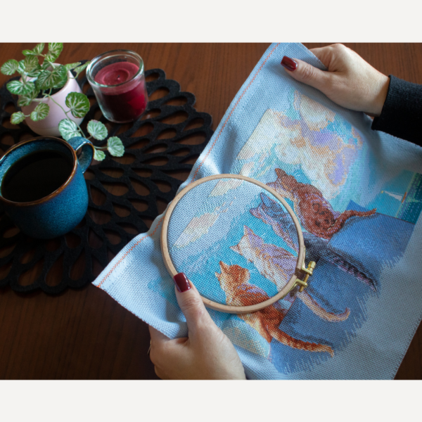 A completed cross-stitch landscape showing four cats (orange, cream, dark grey, and brown) sitting on a blue wall with their backs to the viewer. They are looking out over a blue ocean with a small white sailboat, under a sky filled with fluffy yellow and pink clouds.