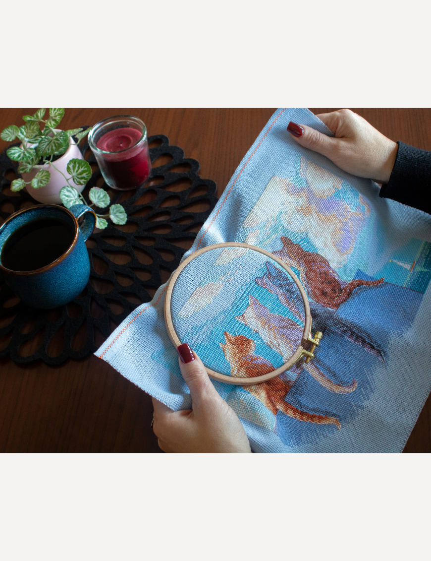 A completed cross-stitch landscape showing four cats (orange, cream, dark grey, and brown) sitting on a blue wall with their backs to the viewer. They are looking out over a blue ocean with a small white sailboat, under a sky filled with fluffy yellow and pink clouds.