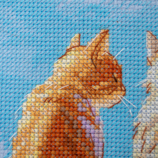 A completed cross-stitch landscape showing four cats (orange, cream, dark grey, and brown) sitting on a blue wall with their backs to the viewer. They are looking out over a blue ocean with a small white sailboat, under a sky filled with fluffy yellow and pink clouds.