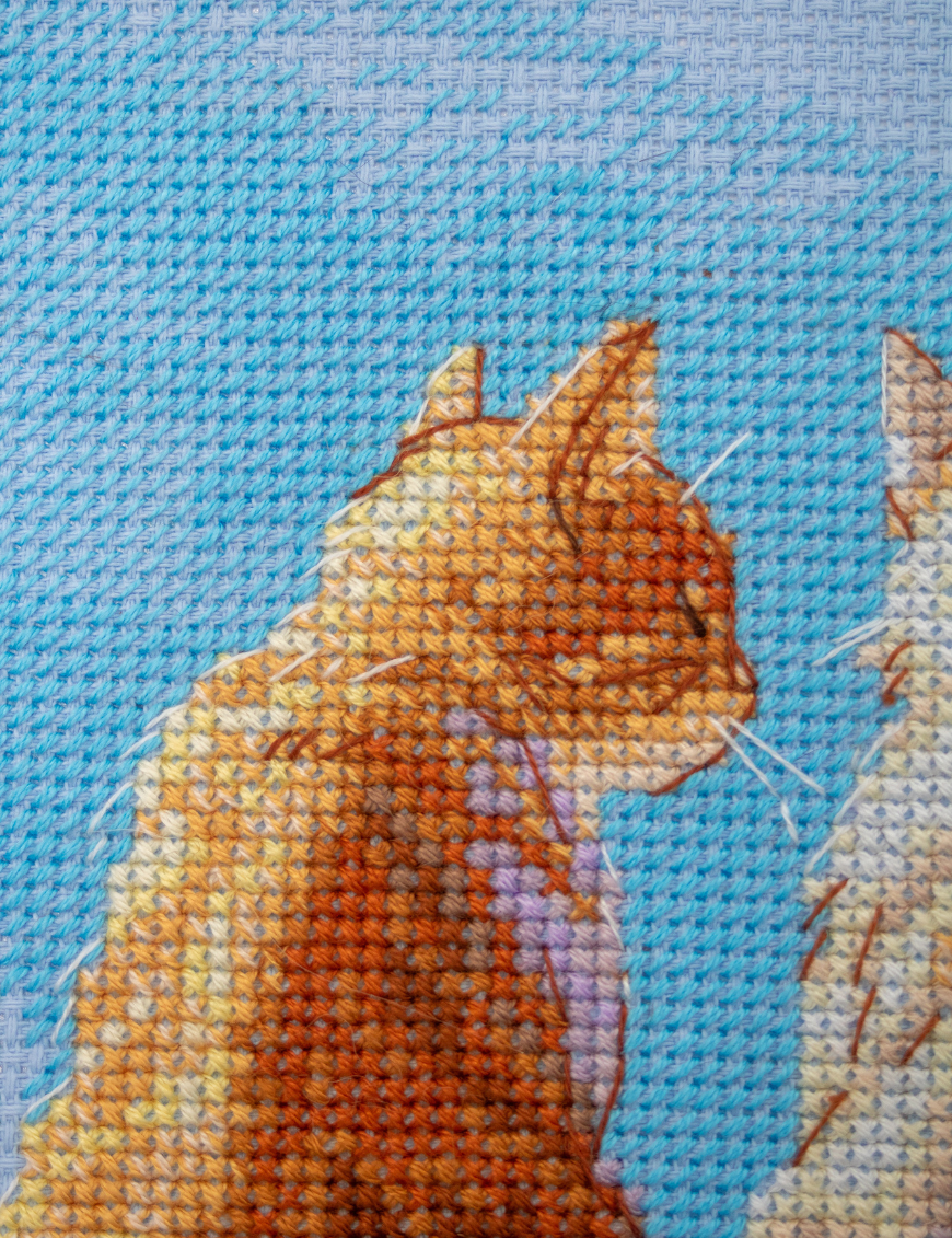 A completed cross-stitch landscape showing four cats (orange, cream, dark grey, and brown) sitting on a blue wall with their backs to the viewer. They are looking out over a blue ocean with a small white sailboat, under a sky filled with fluffy yellow and pink clouds.
