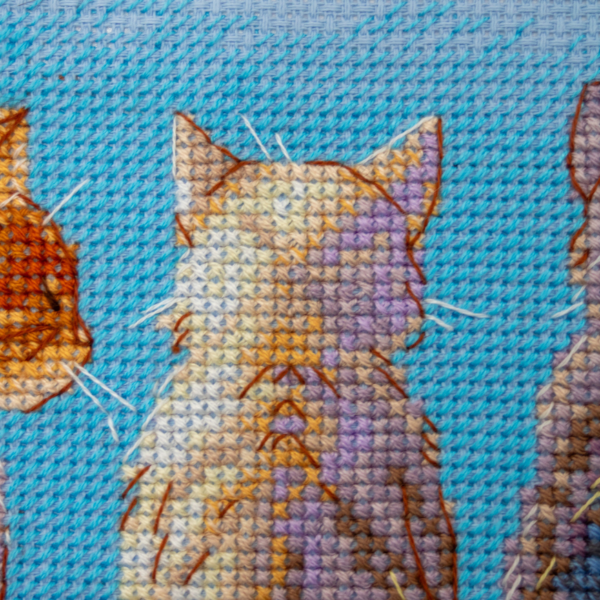 A completed cross-stitch landscape showing four cats (orange, cream, dark grey, and brown) sitting on a blue wall with their backs to the viewer. They are looking out over a blue ocean with a small white sailboat, under a sky filled with fluffy yellow and pink clouds.