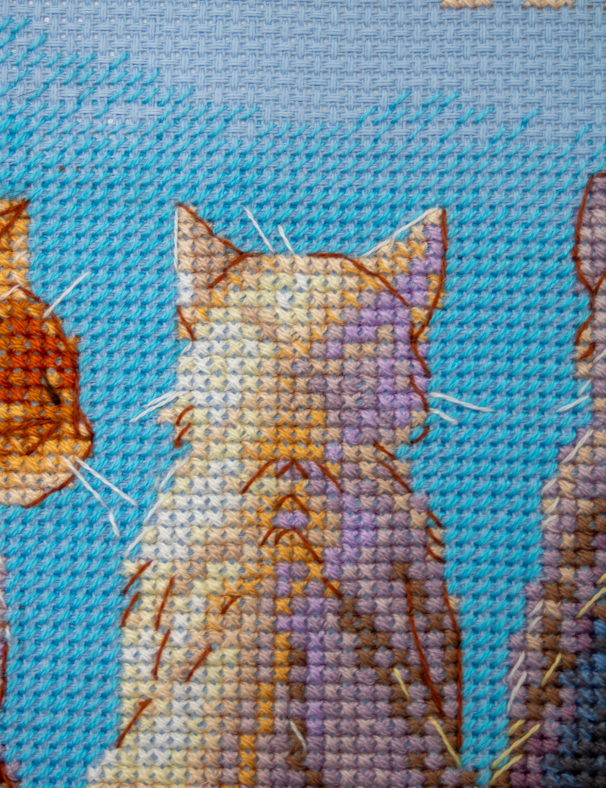 A completed cross-stitch landscape showing four cats (orange, cream, dark grey, and brown) sitting on a blue wall with their backs to the viewer. They are looking out over a blue ocean with a small white sailboat, under a sky filled with fluffy yellow and pink clouds.