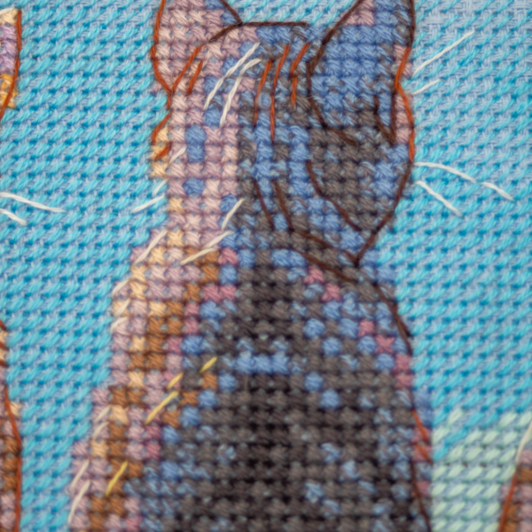 A completed cross-stitch landscape showing four cats (orange, cream, dark grey, and brown) sitting on a blue wall with their backs to the viewer. They are looking out over a blue ocean with a small white sailboat, under a sky filled with fluffy yellow and pink clouds.