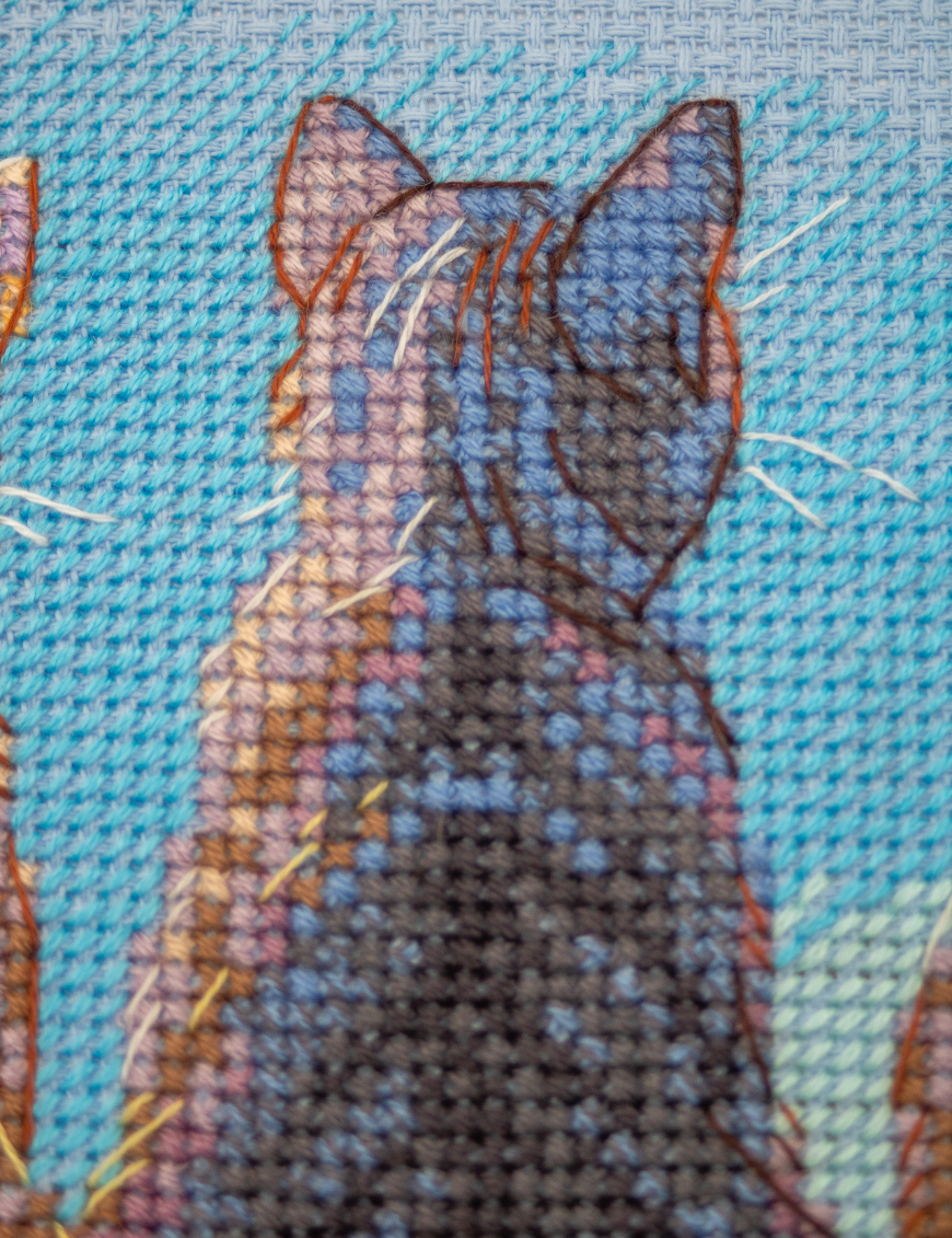 A completed cross-stitch landscape showing four cats (orange, cream, dark grey, and brown) sitting on a blue wall with their backs to the viewer. They are looking out over a blue ocean with a small white sailboat, under a sky filled with fluffy yellow and pink clouds.