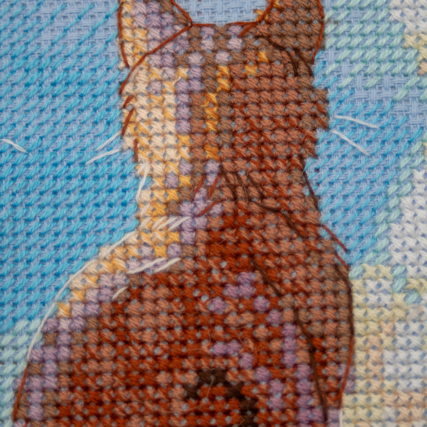 A completed cross-stitch landscape showing four cats (orange, cream, dark grey, and brown) sitting on a blue wall with their backs to the viewer. They are looking out over a blue ocean with a small white sailboat, under a sky filled with fluffy yellow and pink clouds.
