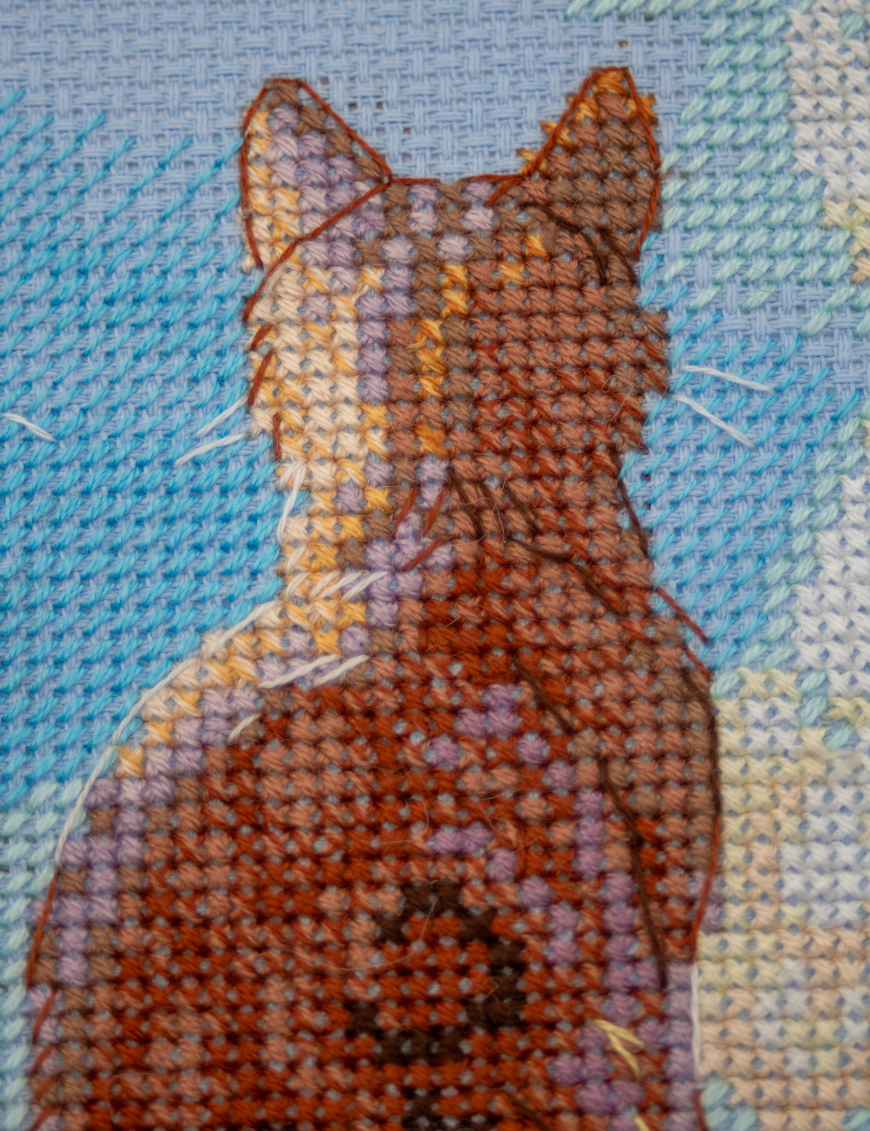 A completed cross-stitch landscape showing four cats (orange, cream, dark grey, and brown) sitting on a blue wall with their backs to the viewer. They are looking out over a blue ocean with a small white sailboat, under a sky filled with fluffy yellow and pink clouds.