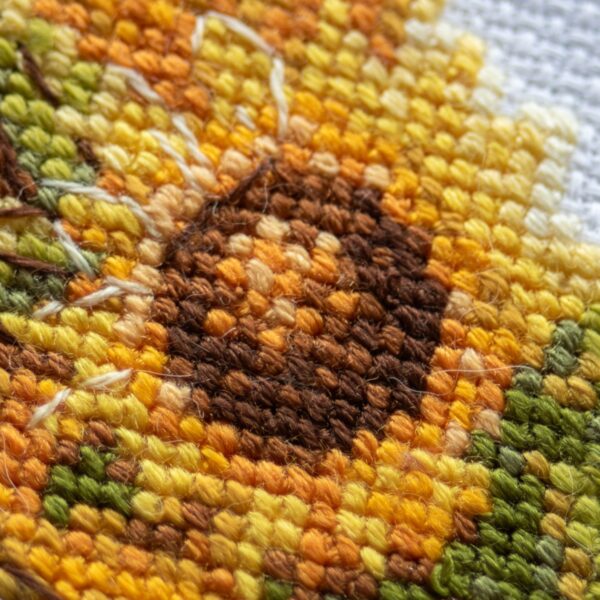 A1043 Sunshine Gnome Aine Cross-Stitch Kit featuring a garden gnome in a yellow sunflower hat surrounded by red flowers, daisies, and green foliage.
