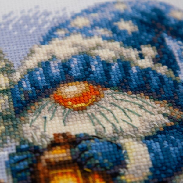 A1045 Snowy Gnome Aine Cross-Stitch Kit featuring a winter gnome with a blue snowy hat, glowing lantern, wooden sled, bird, and snow-covered evergreen trees.