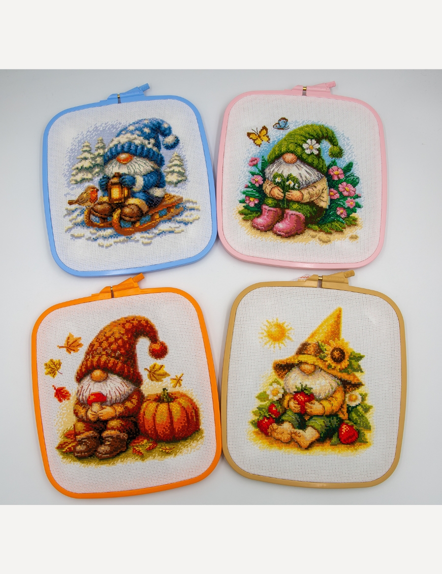 Four seasonal Aine gnome cross-stitch designs displayed in colorful square embroidery hoops, featuring Blossom, Sunshine, Harvest, and Snowy Gnomes.