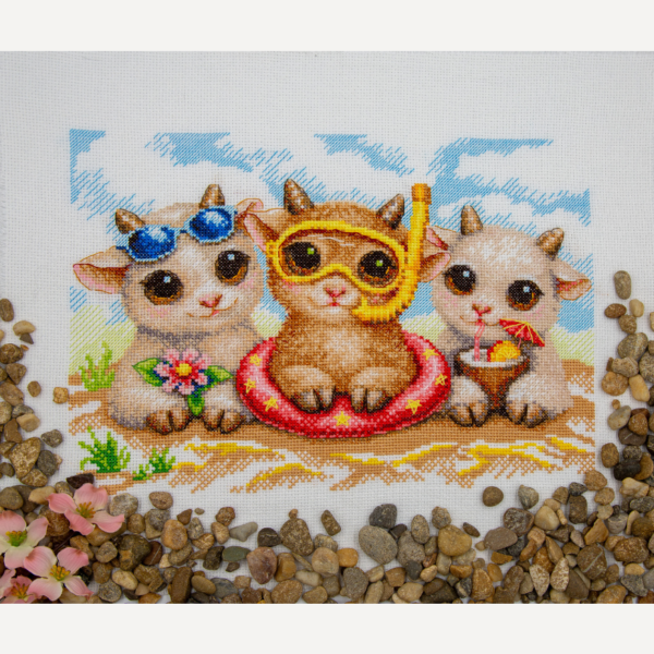 A completed cross-stitch picture of three baby goats on a beach: one wearing blue sunglasses beside a pink flower, one wearing a yellow snorkel mask and tube inside a red inflatable swim ring with yellow stars, and one drinking a tropical cocktail with a paper umbrella, set against a blue sky with white clouds.