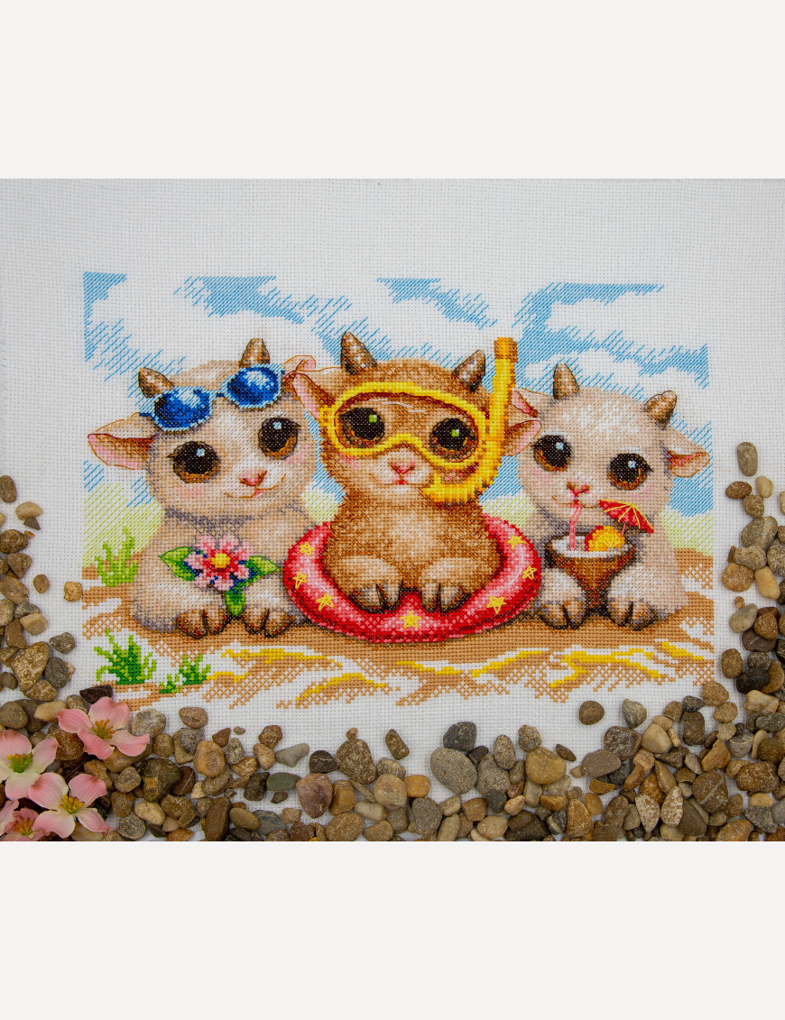 A completed cross-stitch picture of three baby goats on a beach: one wearing blue sunglasses beside a pink flower, one wearing a yellow snorkel mask and tube inside a red inflatable swim ring with yellow stars, and one drinking a tropical cocktail with a paper umbrella, set against a blue sky with white clouds.