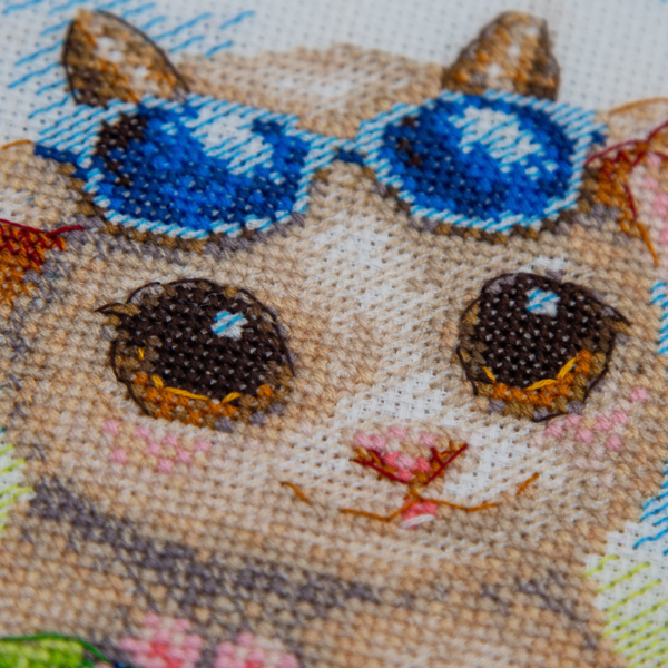 A completed cross-stitch picture of three baby goats on a beach: one wearing blue sunglasses beside a pink flower, one wearing a yellow snorkel mask and tube inside a red inflatable swim ring with yellow stars, and one drinking a tropical cocktail with a paper umbrella, set against a blue sky with white clouds.