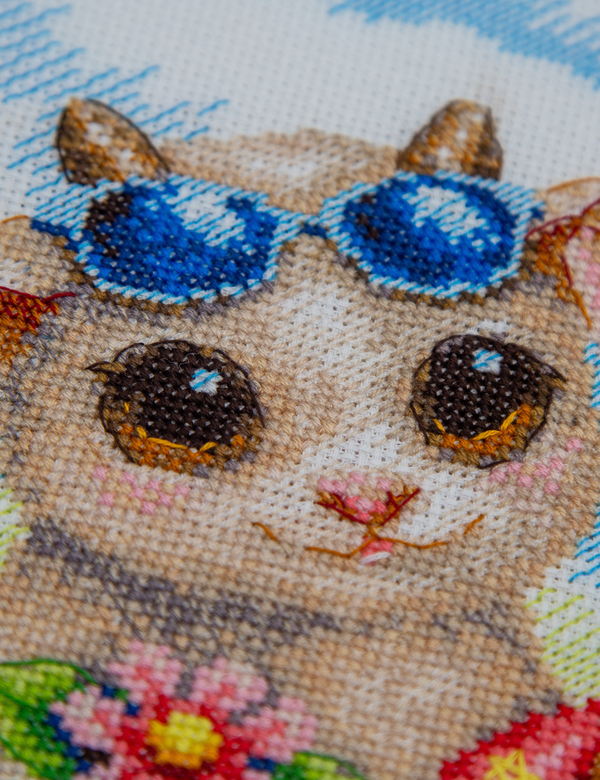 A completed cross-stitch picture of three baby goats on a beach: one wearing blue sunglasses beside a pink flower, one wearing a yellow snorkel mask and tube inside a red inflatable swim ring with yellow stars, and one drinking a tropical cocktail with a paper umbrella, set against a blue sky with white clouds.
