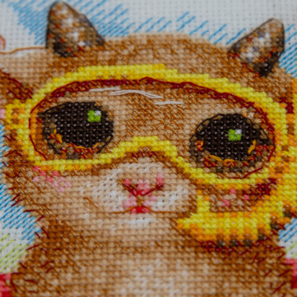 A completed cross-stitch picture of three baby goats on a beach: one wearing blue sunglasses beside a pink flower, one wearing a yellow snorkel mask and tube inside a red inflatable swim ring with yellow stars, and one drinking a tropical cocktail with a paper umbrella, set against a blue sky with white clouds.