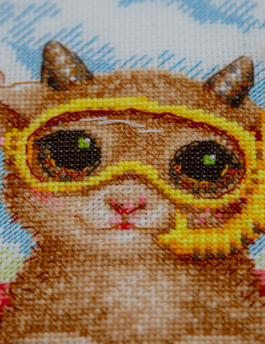 A completed cross-stitch picture of three baby goats on a beach: one wearing blue sunglasses beside a pink flower, one wearing a yellow snorkel mask and tube inside a red inflatable swim ring with yellow stars, and one drinking a tropical cocktail with a paper umbrella, set against a blue sky with white clouds.