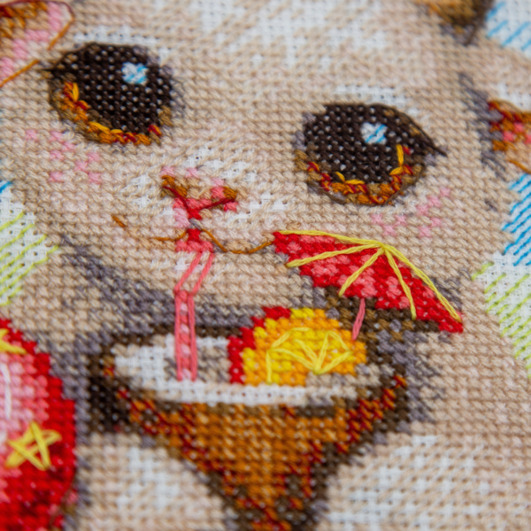 A completed cross-stitch picture of three baby goats on a beach: one wearing blue sunglasses beside a pink flower, one wearing a yellow snorkel mask and tube inside a red inflatable swim ring with yellow stars, and one drinking a tropical cocktail with a paper umbrella, set against a blue sky with white clouds.