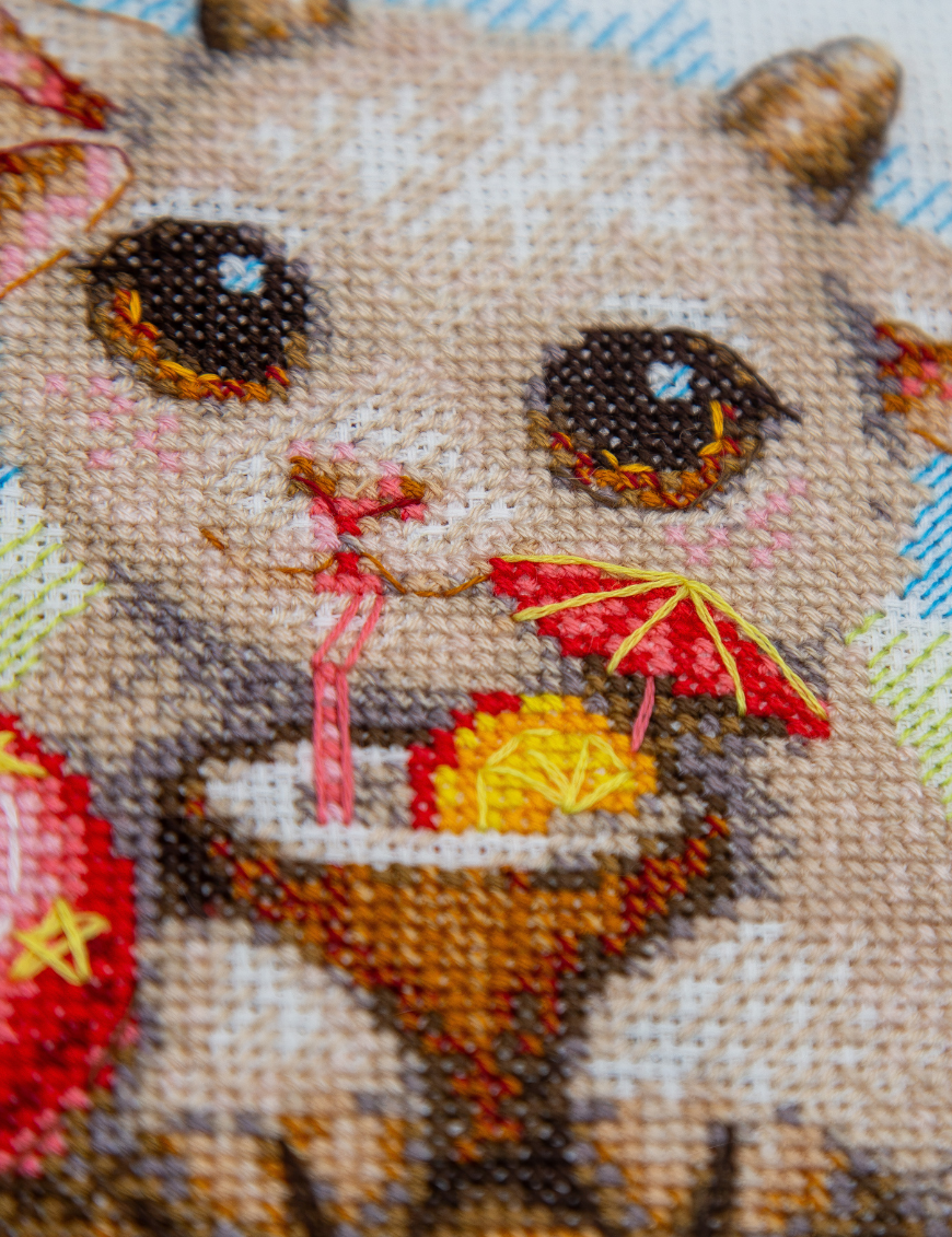 A completed cross-stitch picture of three baby goats on a beach: one wearing blue sunglasses beside a pink flower, one wearing a yellow snorkel mask and tube inside a red inflatable swim ring with yellow stars, and one drinking a tropical cocktail with a paper umbrella, set against a blue sky with white clouds.