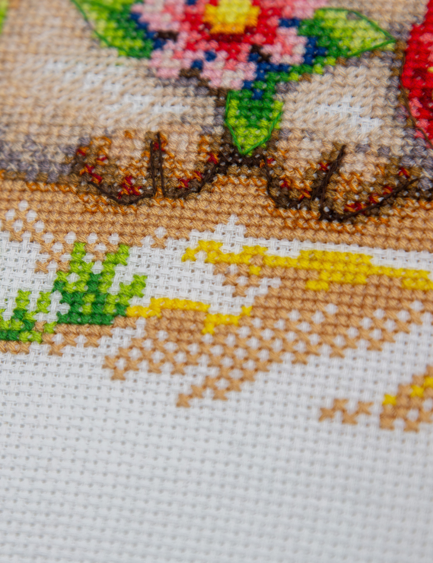 A completed cross-stitch picture of three baby goats on a beach: one wearing blue sunglasses beside a pink flower, one wearing a yellow snorkel mask and tube inside a red inflatable swim ring with yellow stars, and one drinking a tropical cocktail with a paper umbrella, set against a blue sky with white clouds.