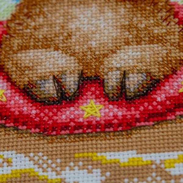 A completed cross-stitch picture of three baby goats on a beach: one wearing blue sunglasses beside a pink flower, one wearing a yellow snorkel mask and tube inside a red inflatable swim ring with yellow stars, and one drinking a tropical cocktail with a paper umbrella, set against a blue sky with white clouds.