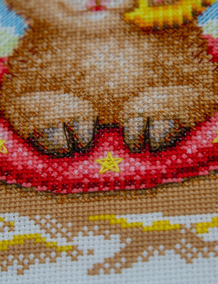 A completed cross-stitch picture of three baby goats on a beach: one wearing blue sunglasses beside a pink flower, one wearing a yellow snorkel mask and tube inside a red inflatable swim ring with yellow stars, and one drinking a tropical cocktail with a paper umbrella, set against a blue sky with white clouds.