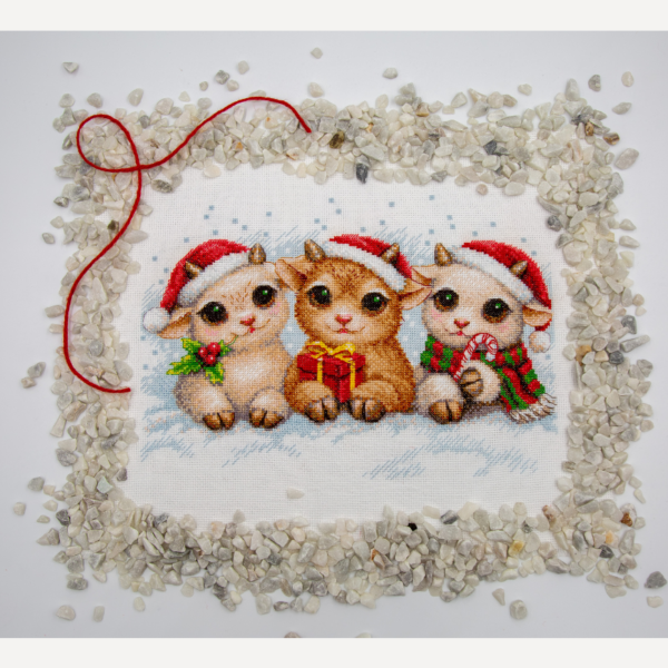 A completed cross-stitch picture of three baby goats wearing red Santa hats, huddled together in falling snow: one with a sprig of holly and red berries, one holding a wrapped Christmas present with a red bow, and one wearing a red-and-green plaid scarf and holding a candy cane.