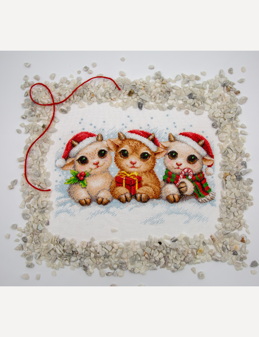 A completed cross-stitch picture of three baby goats wearing red Santa hats, huddled together in falling snow: one with a sprig of holly and red berries, one holding a wrapped Christmas present with a red bow, and one wearing a red-and-green plaid scarf and holding a candy cane.