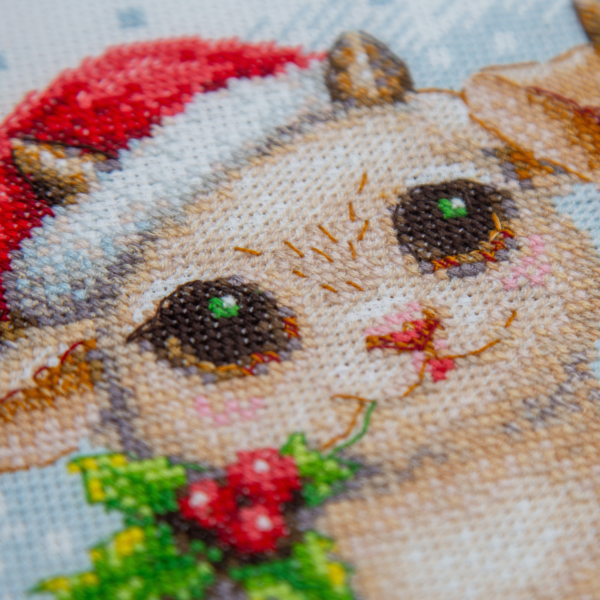 A completed cross-stitch picture of three baby goats wearing red Santa hats, huddled together in falling snow: one with a sprig of holly and red berries, one holding a wrapped Christmas present with a red bow, and one wearing a red-and-green plaid scarf and holding a candy cane.