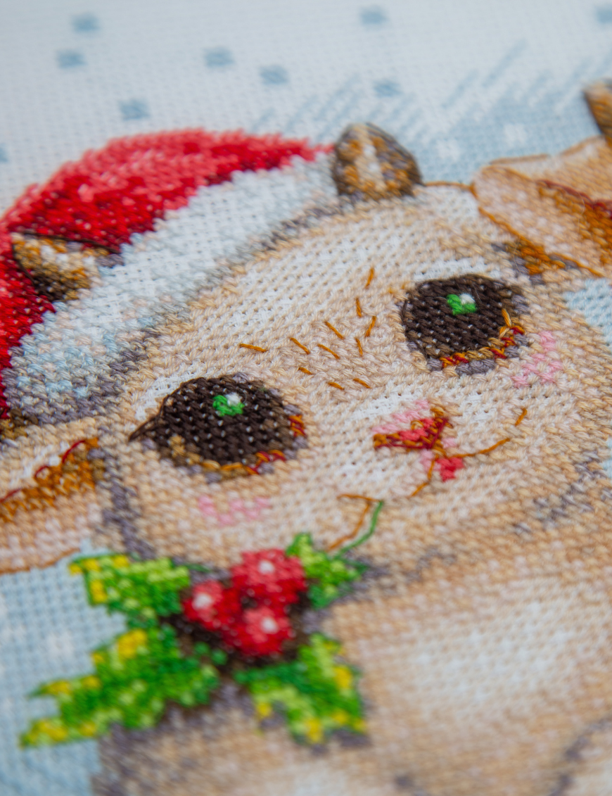 A completed cross-stitch picture of three baby goats wearing red Santa hats, huddled together in falling snow: one with a sprig of holly and red berries, one holding a wrapped Christmas present with a red bow, and one wearing a red-and-green plaid scarf and holding a candy cane.