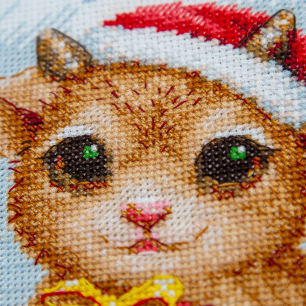 A completed cross-stitch picture of three baby goats wearing red Santa hats, huddled together in falling snow: one with a sprig of holly and red berries, one holding a wrapped Christmas present with a red bow, and one wearing a red-and-green plaid scarf and holding a candy cane.