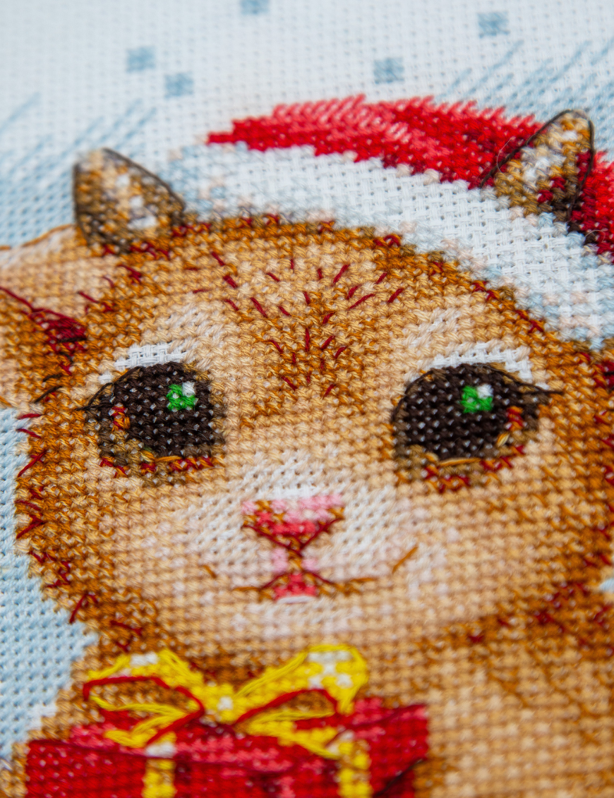 A completed cross-stitch picture of three baby goats wearing red Santa hats, huddled together in falling snow: one with a sprig of holly and red berries, one holding a wrapped Christmas present with a red bow, and one wearing a red-and-green plaid scarf and holding a candy cane.