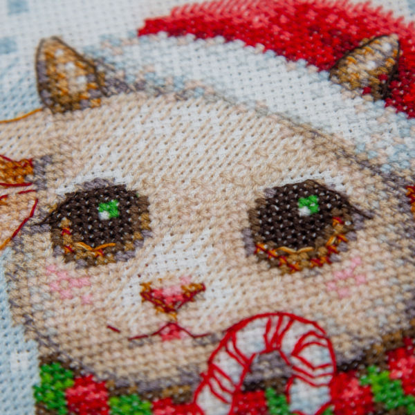 A completed cross-stitch picture of three baby goats wearing red Santa hats, huddled together in falling snow: one with a sprig of holly and red berries, one holding a wrapped Christmas present with a red bow, and one wearing a red-and-green plaid scarf and holding a candy cane.