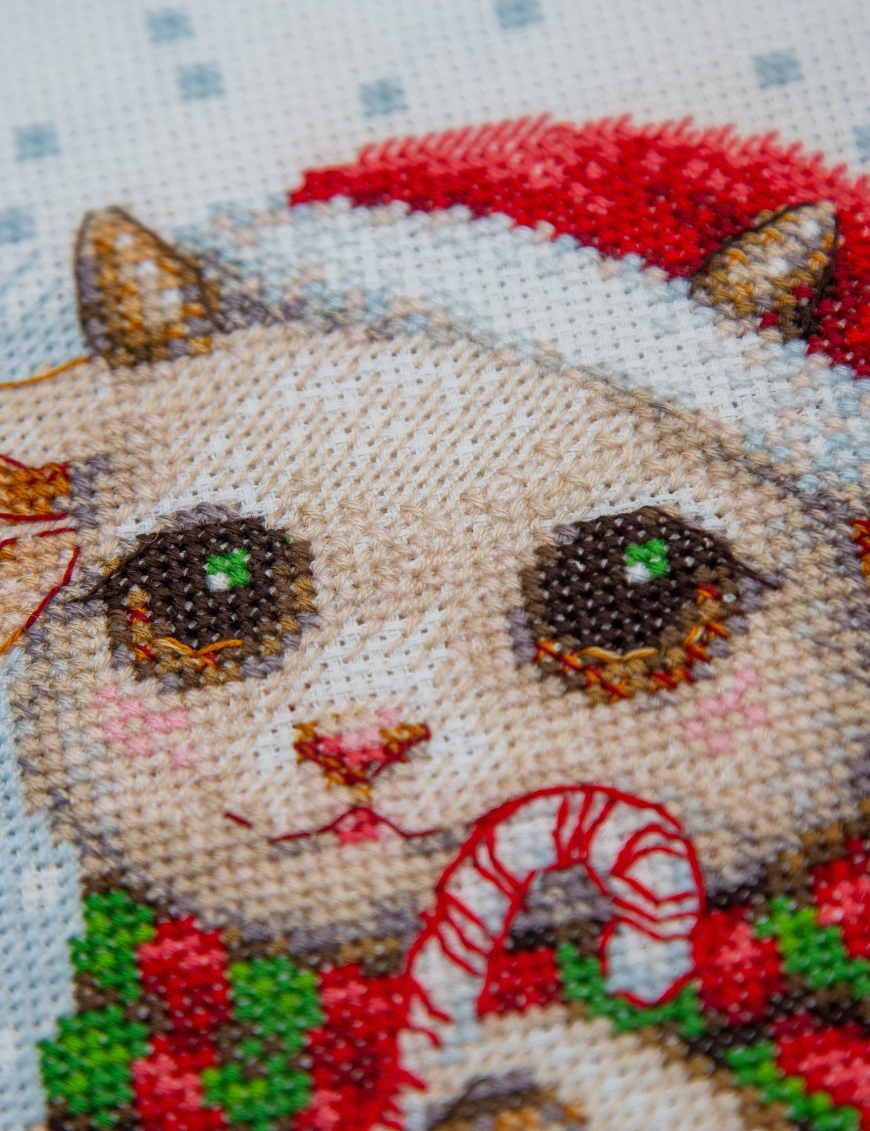 A completed cross-stitch picture of three baby goats wearing red Santa hats, huddled together in falling snow: one with a sprig of holly and red berries, one holding a wrapped Christmas present with a red bow, and one wearing a red-and-green plaid scarf and holding a candy cane.