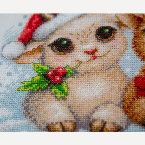 A completed cross-stitch picture of three baby goats wearing red Santa hats, huddled together in falling snow: one with a sprig of holly and red berries, one holding a wrapped Christmas present with a red bow, and one wearing a red-and-green plaid scarf and holding a candy cane.
